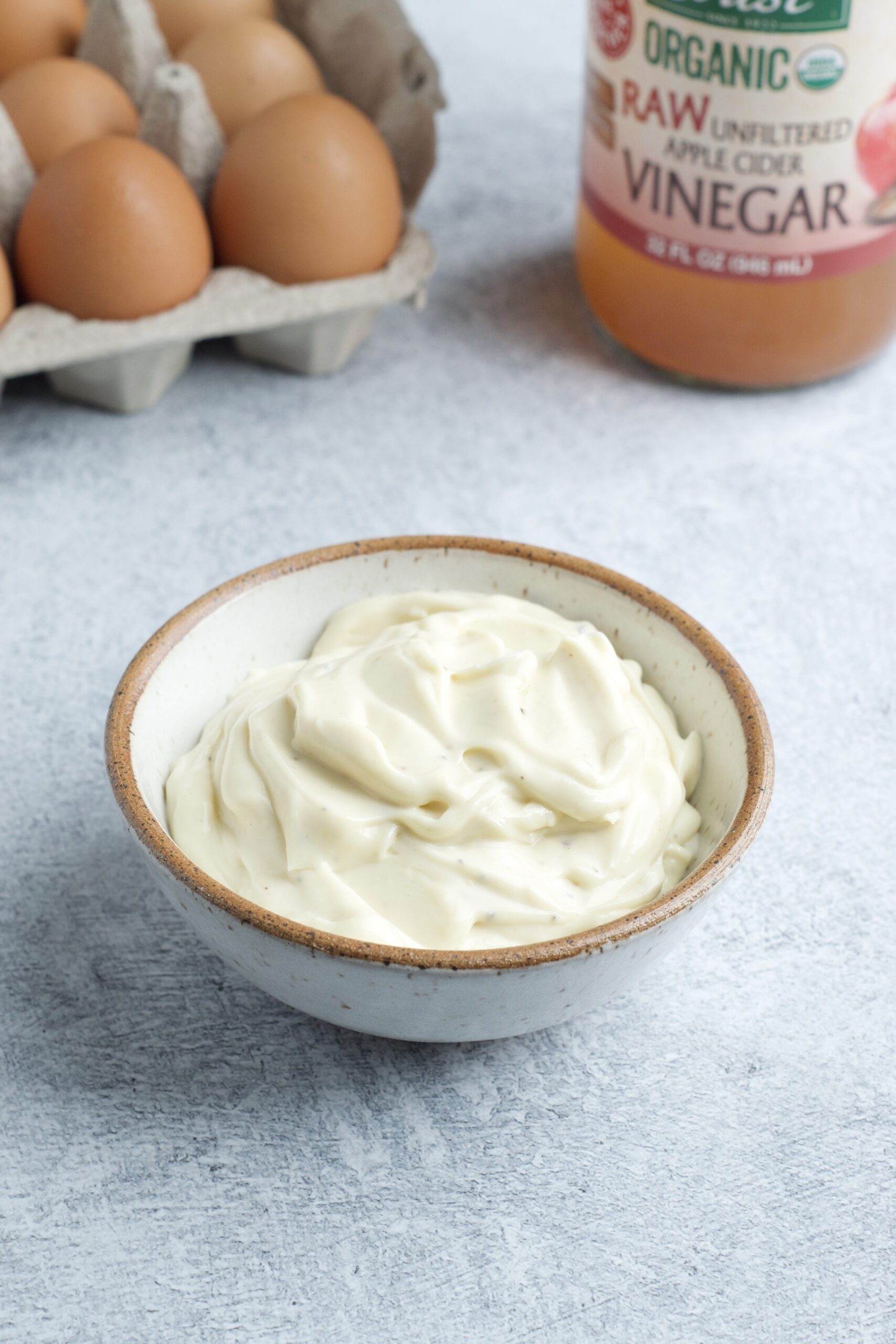 Small bowl of mayo with a carton of eggs and a bottle of amber liquid behind it